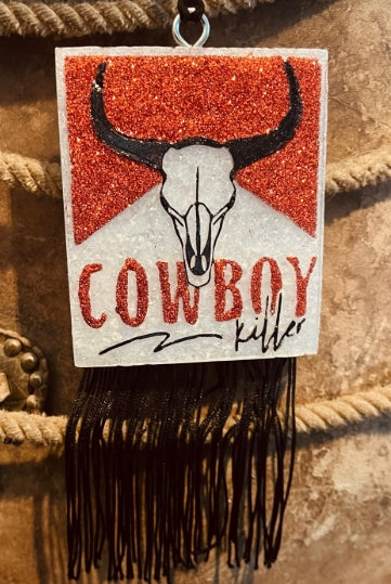 Decorative sign with a bull skull and 'Cowboy Killer' text on a red glittery background, hanging against a rustic background.