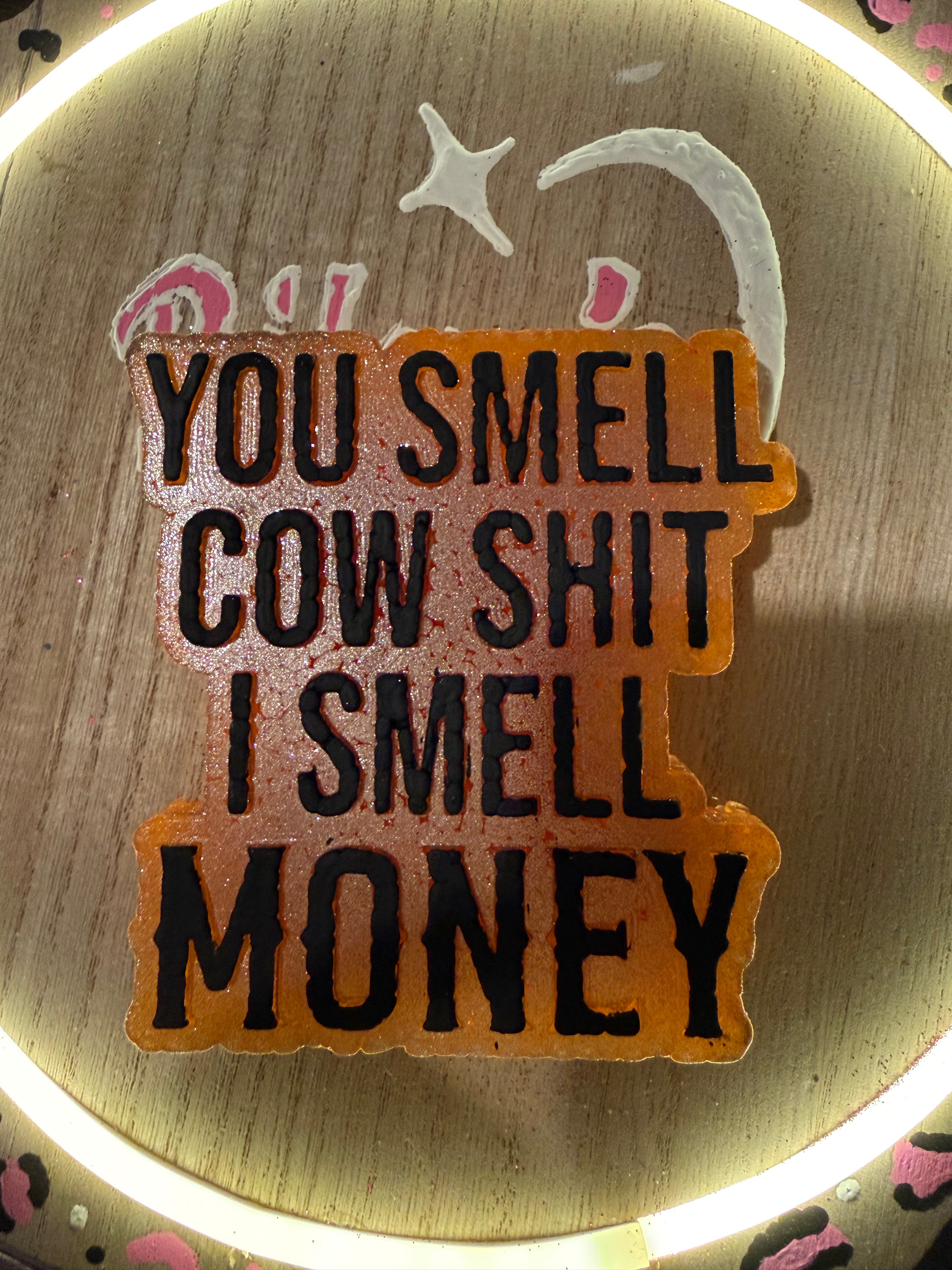 Wooden sign with humorous text 'You smell cow shit, I smell money' on a wooden background.
