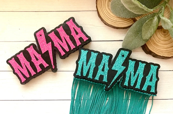 Two patches with 'MAMA' text, one in pink and black, the other in teal and black, on a wooden surface.